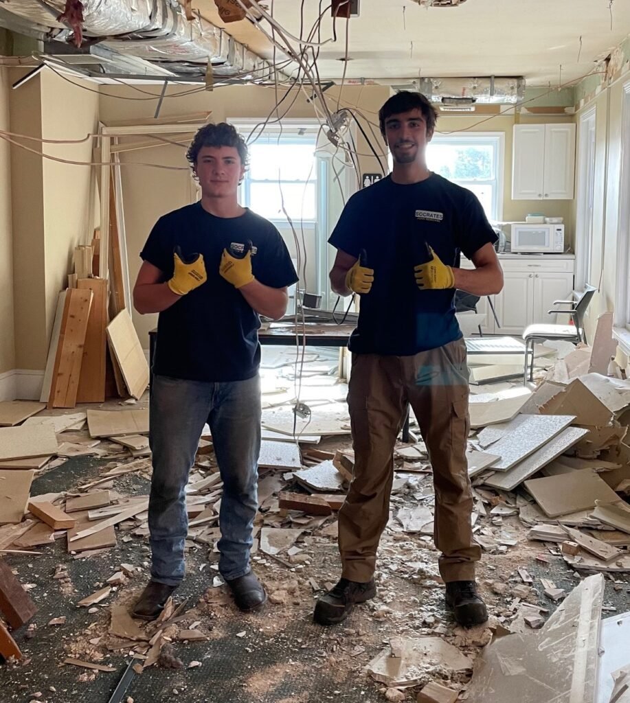 Socrates Junk Removal team performing interior demolition, standing in a debris-filled room with exposed wiring and removed walls in the Wayne area